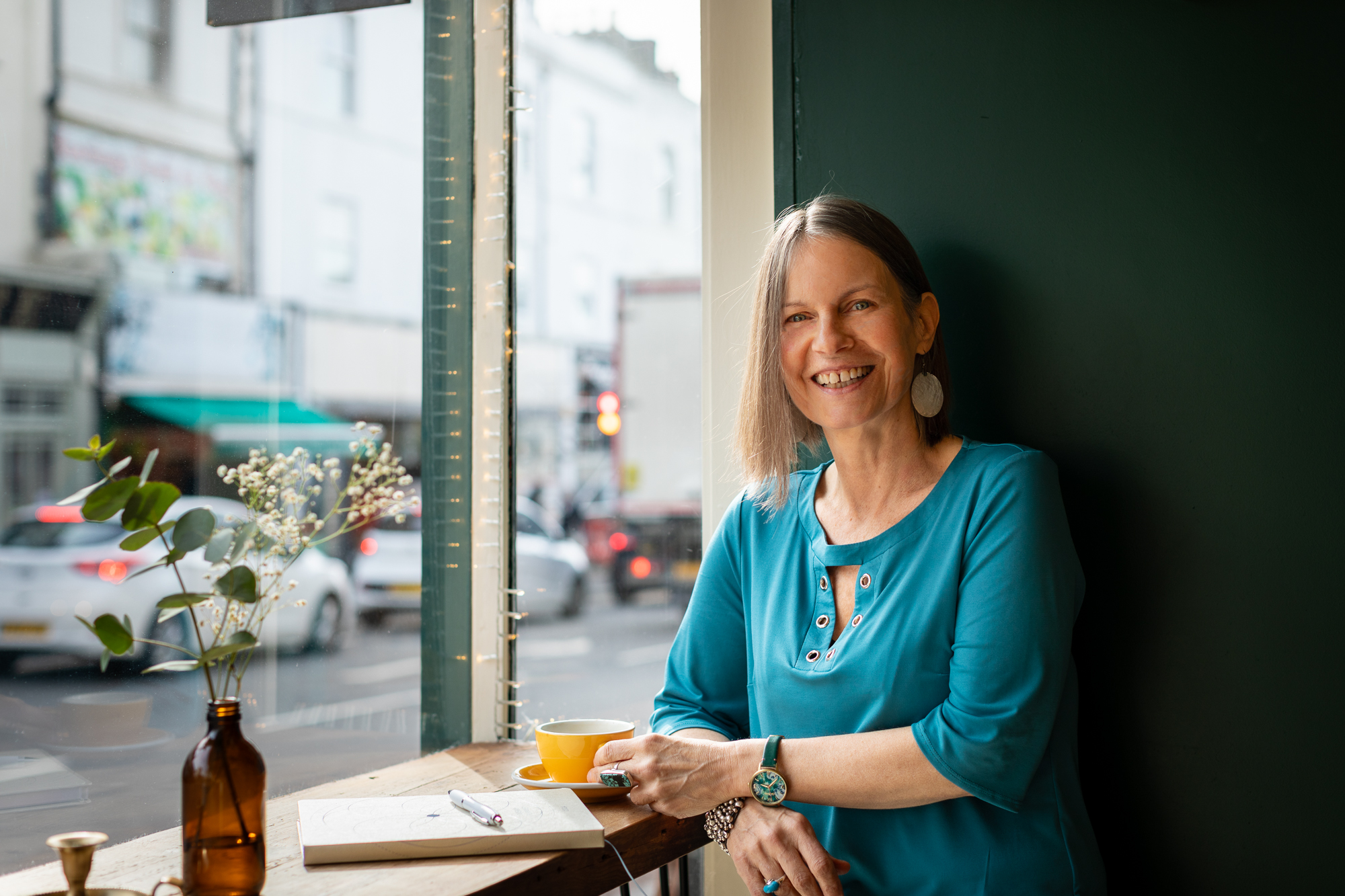 Nicky smiling in a cafe window seat, holding a yellow cup